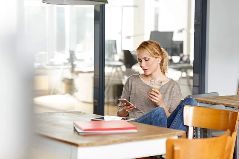 Frau hält ein Latte Macchiato und schaut in einem modernen Büro auf ihr Smartphone